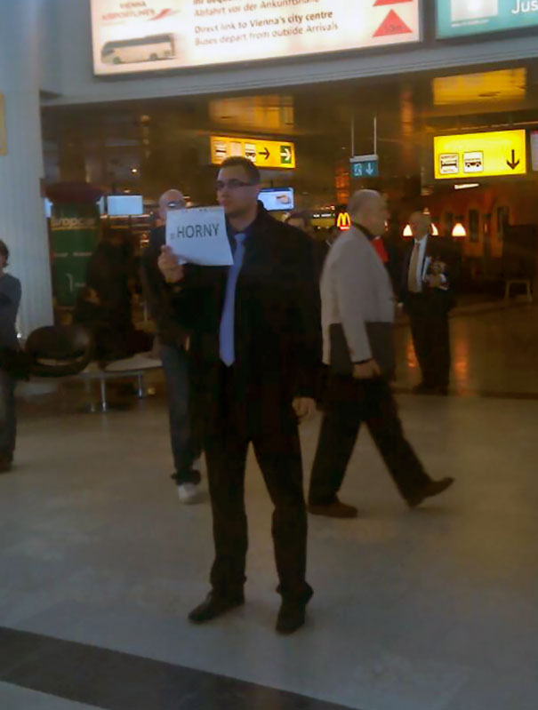 Man at airport holding a funny pickup sign that reads "H***y" amidst a busy terminal.