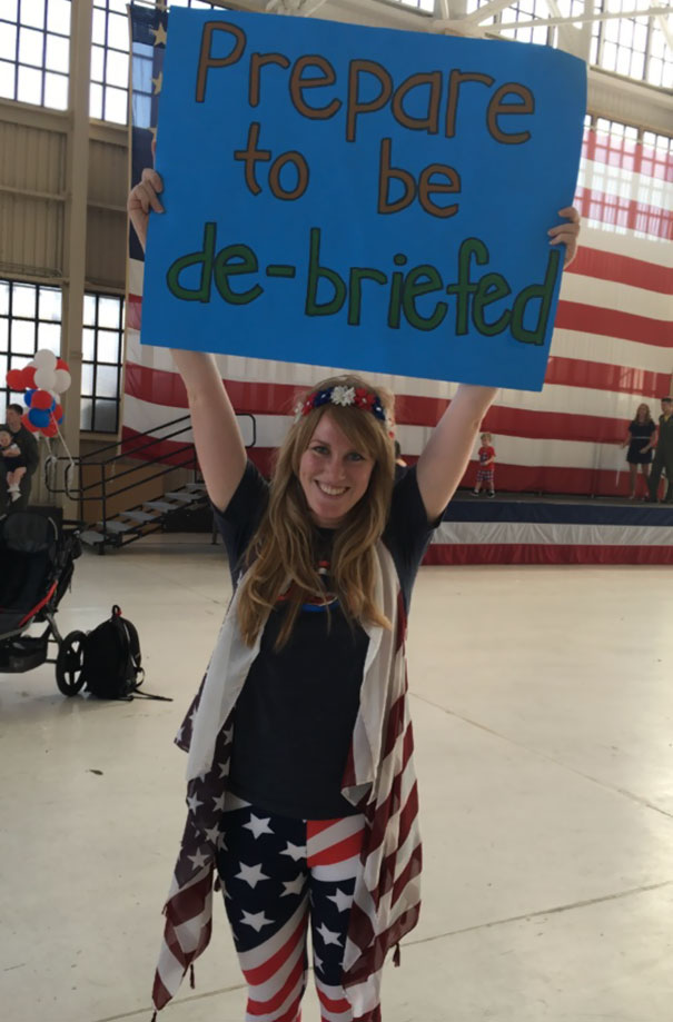 Woman holding funny airport pickup sign saying "Prepare to be de-briefed" in a patriotic outfit.