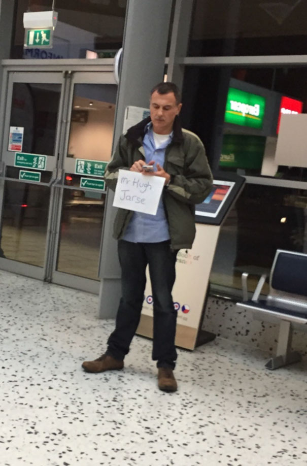 Man at airport holding funny sign that says "Mr. Hugh Jarse," standing near an exit.