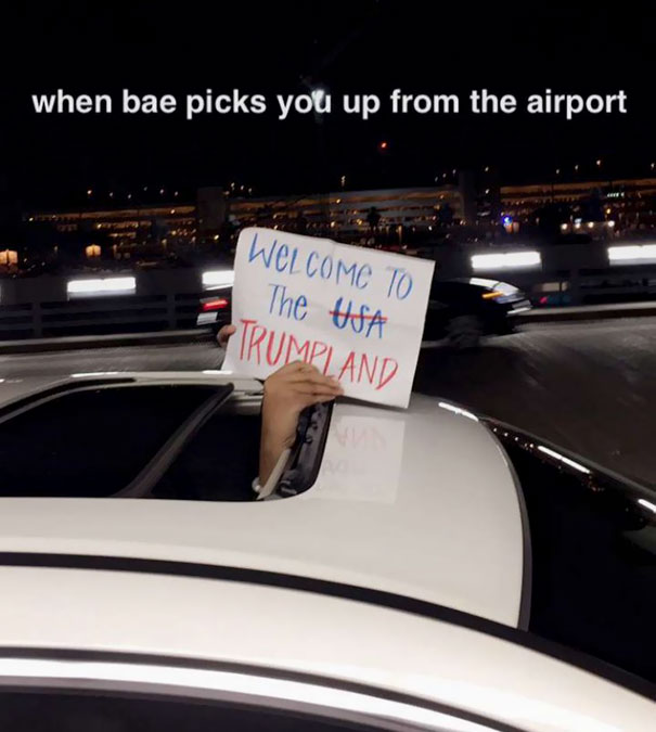 Funny airport pickup sign reading "Welcome to Trumpland" held through car sunroof at night.