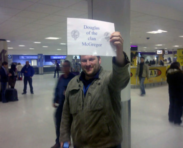 Man at airport holding a funny pickup sign for "Douglas of the clan McGregor," making travelers chuckle.