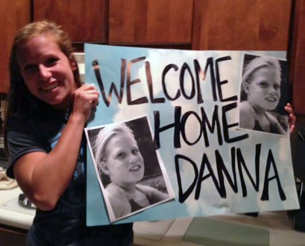 Woman holding a funny airport pickup sign with photos and "Welcome Home Danna" text.