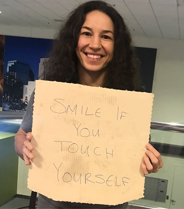 Woman holding a funny airport pickup sign, smiling indoors.