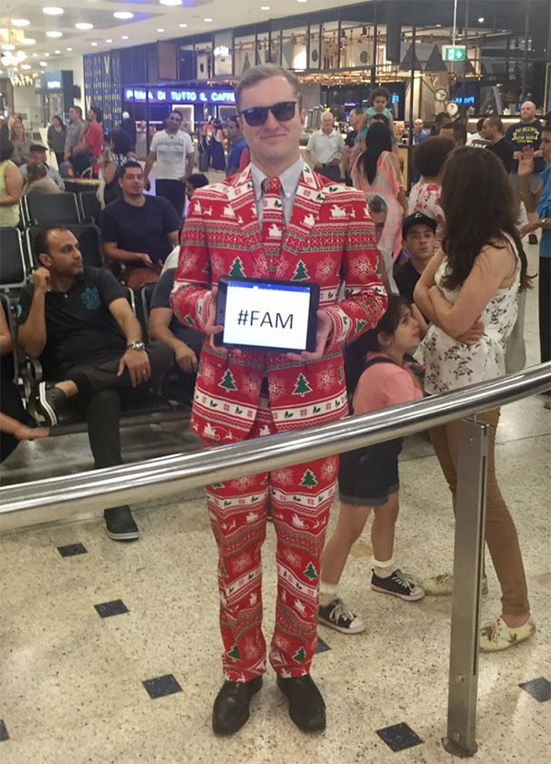 Man in festive suit at airport holding a funny pickup sign with "#FAM," making the crowd chuckle.
