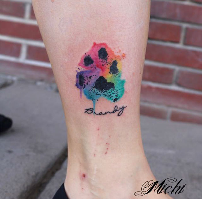 Colorful dog paw print tattoo with the name "Brandy" on a person's leg.