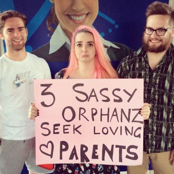 Group holding a funny airport pickup sign reading "3 Sassy Orphanz Seek Loving Parents" in colorful attire.