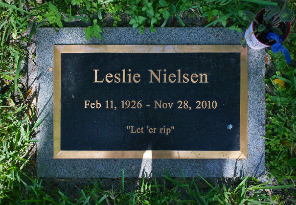 Leslie Nielsen tombstone with humorous epitaph Let 'er rip surrounded by grass and small potted plant outdoors