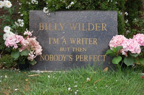 Tombstone engraved with a humorous writer’s epitaph surrounded by pink flowers and green grass in a cemetery setting.