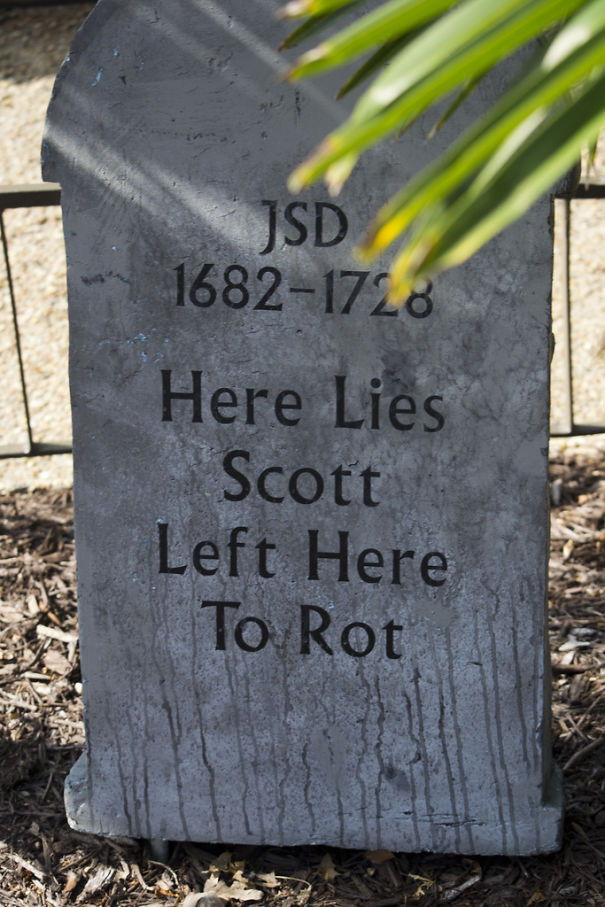 Old gray tombstone with a funny epitaph about Scott left to rot, showcasing humor on tombstones in a sunny cemetery setting.
