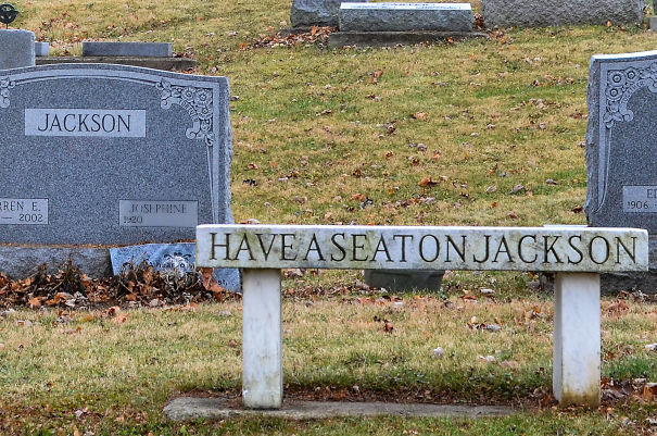 A humorous tombstone bench engraved with Have a Seat on Jackson in a cemetery with other gravestones nearby.