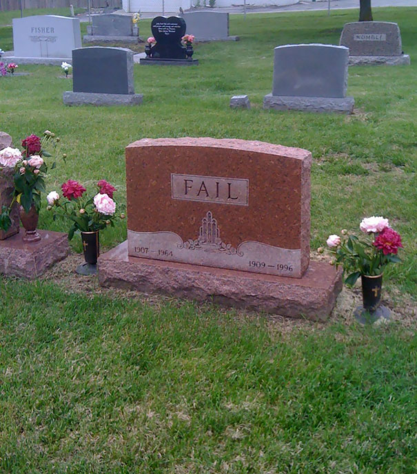 Funny tombstone with the word FAIL engraved, surrounded by flowers on green grass in a cemetery setting.