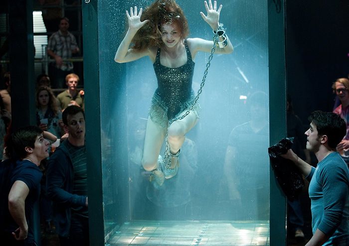 Woman performing an underwater escape illusion in Now You See Me movie, surrounded by an amazed crowd of onlookers.