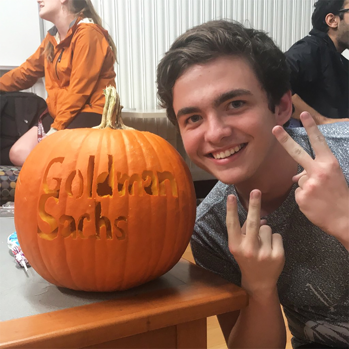 Young person posing with a carved pumpkin displaying "Goldman Sachs."