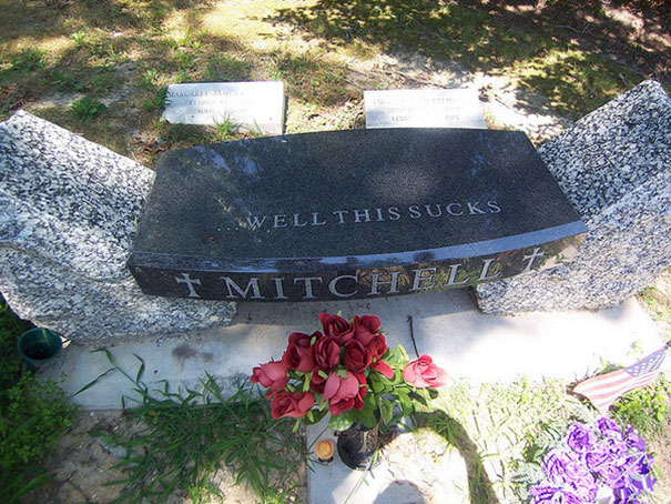 Funny tombstone with the inscription well this s***s on a black granite grave marker surrounded by flowers.