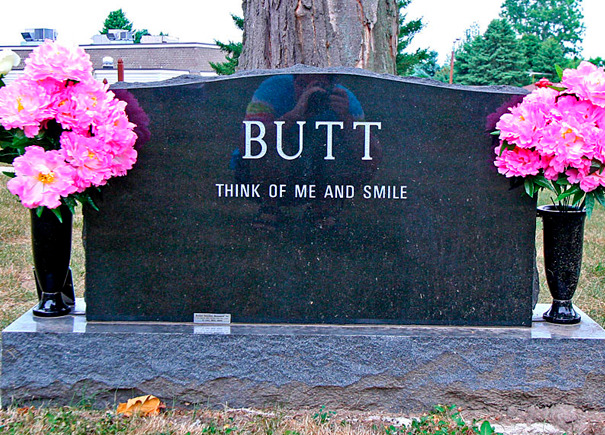 Funny tombstone engraved with the word B**T and the phrase think of me and smile, surrounded by pink flowers.