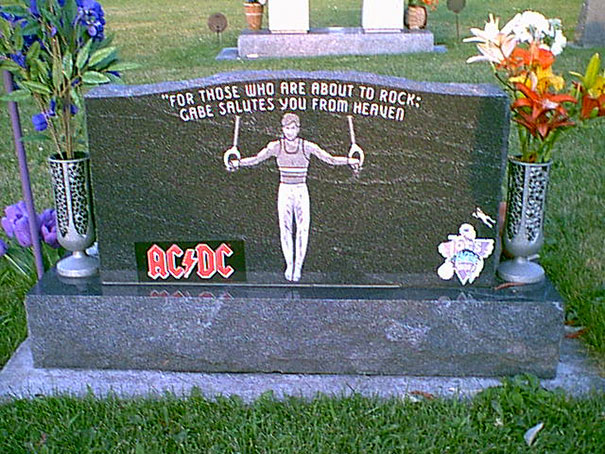 Humorous tombstone with AC/DC tribute and illustration of a gymnast holding rings, surrounded by flowers in a cemetery.