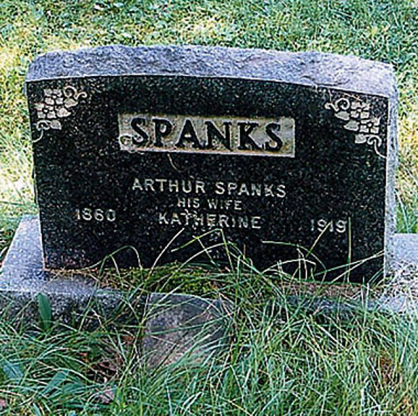 Humorous tombstone of Arthur Spanks and his wife Katherine engraved in black stone with decorative corners outdoors.