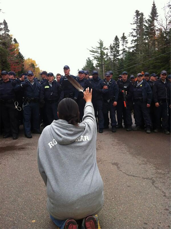 A Woman Kneels Down And Holds Up A Feather While Facing Police Moving In To Break Up Anti-Fracking Protesters In New Brunswick, October 2013