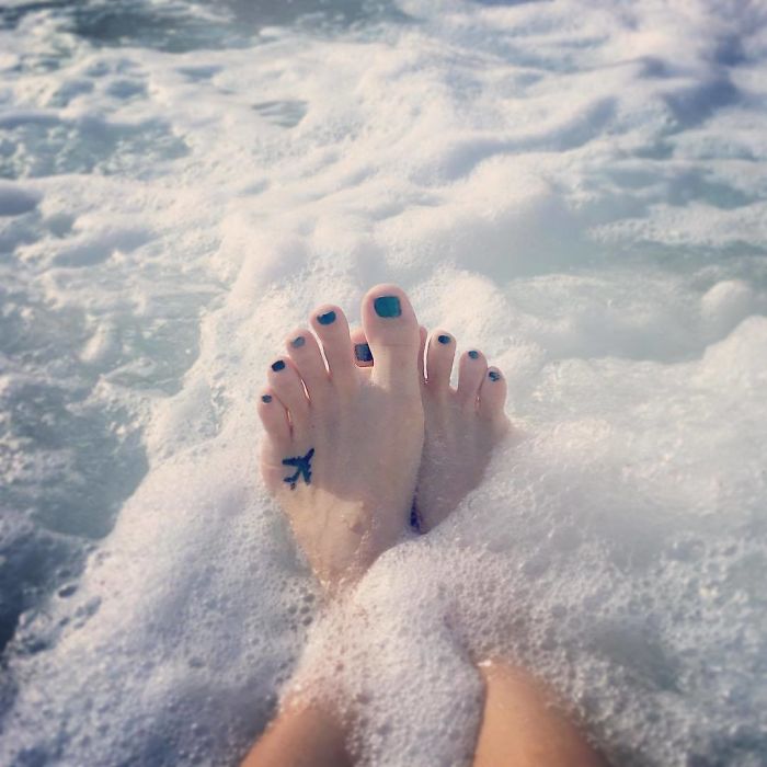 Feet with travel tattoo soaking in ocean waves.
