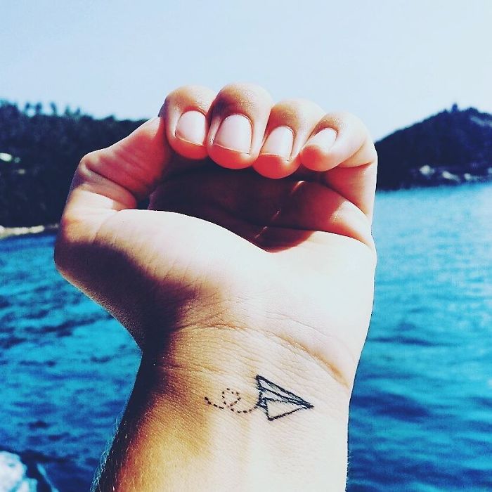 Minimalist travel tattoo of a paper plane on a wrist, with a scenic ocean backdrop, inspiring wanderlust.