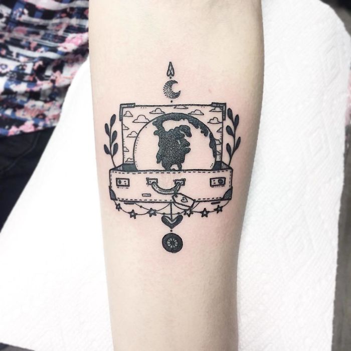 Travel tattoo featuring a suitcase with a globe inside, decorated with stars and plants.