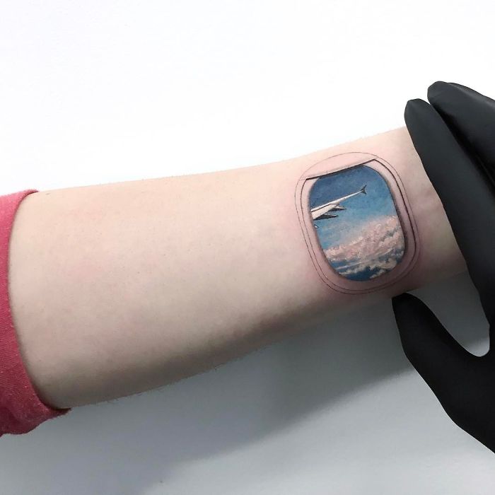 Travel tattoo of an airplane window on arm, featuring clouds and sky, inspiring wanderlust.