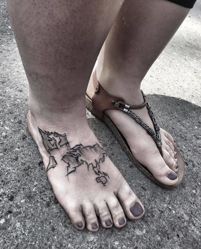Foot with a map tattoo symbolizing travel aspirations, next to a sandal on concrete.