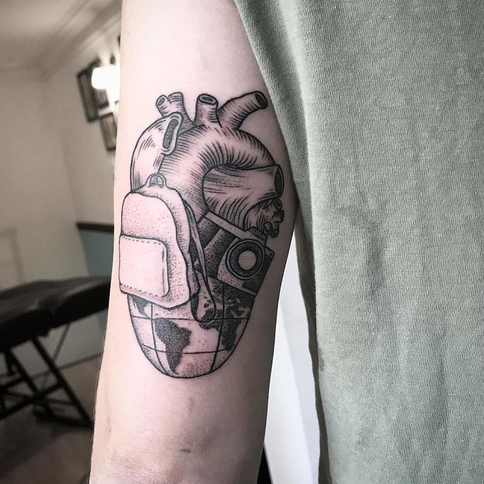 Travel tattoo featuring a world map heart, camera, and backpack on an arm.