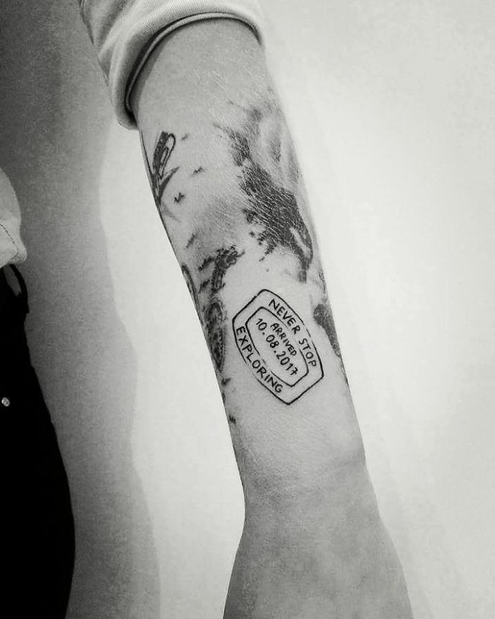Black and white travel tattoo with a passport stamp design on an arm, featuring a motivational quote.