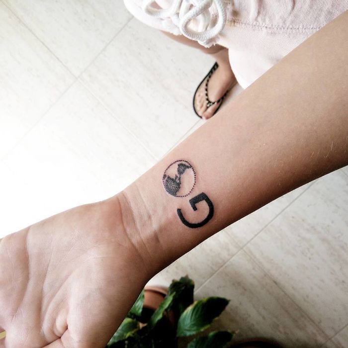 Travel-themed tattoo on a person's wrist featuring a globe and a letter G.