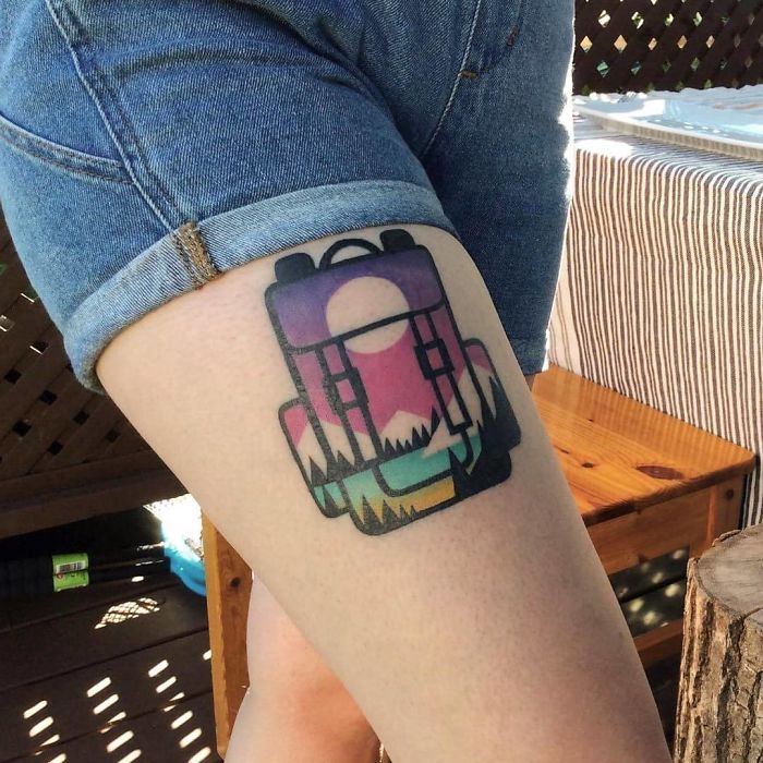 Colorful travel tattoo of a backpack with a sunset and mountains design on a person's thigh.