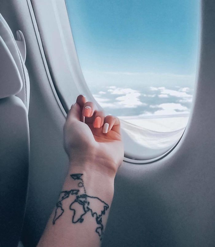 A travel tattoo of a world map on a person's arm, seen by an airplane window overlooking clouds.