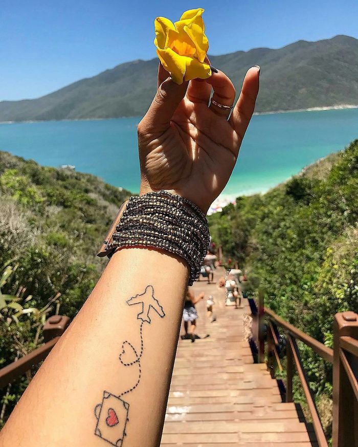 Travel tattoo of an airplane and suitcase on an arm, with a scenic beach view and a yellow flower in hand.