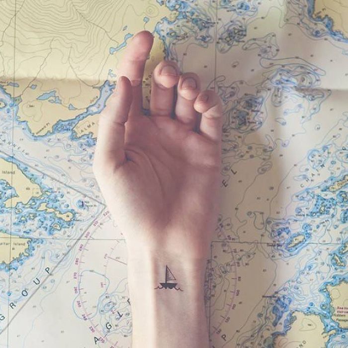 Minimalist travel tattoo of a sailboat on a wrist against a nautical map background.