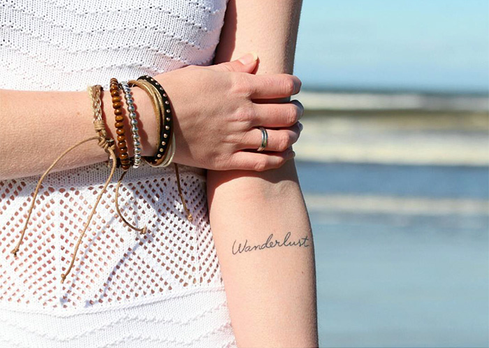 A "Wanderlust" tattoo on a woman's arm by the beach, alongside travel-inspired bracelets.