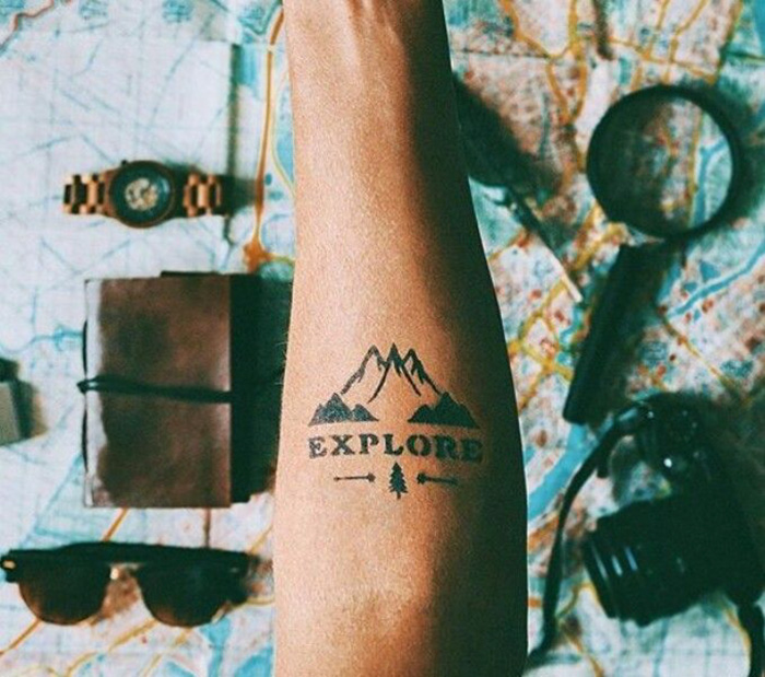 Arm with "Explore" tattoo over a map, surrounded by travel items like a camera, sunglasses, and a notebook.
