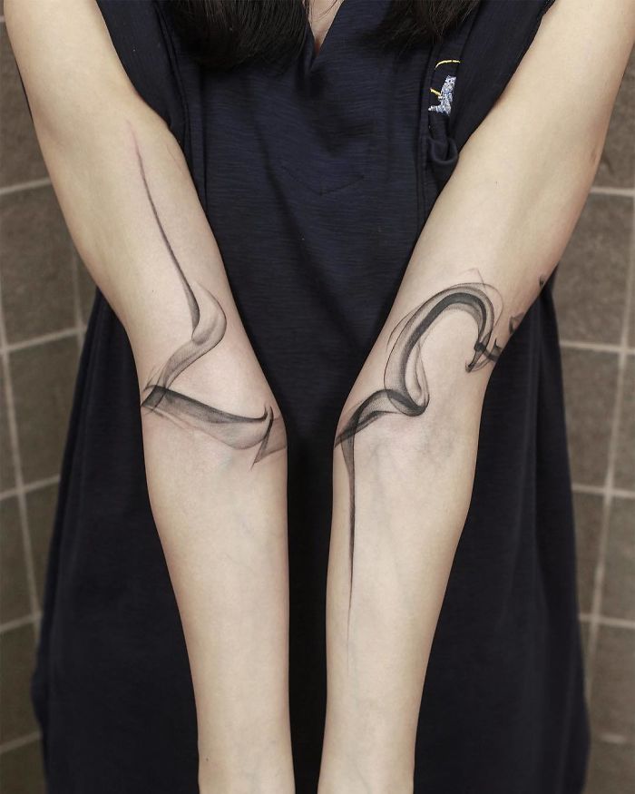 Watercolor tattoos by Chen Jie on forearms, featuring elegant, abstract black ink designs against a neutral background.
