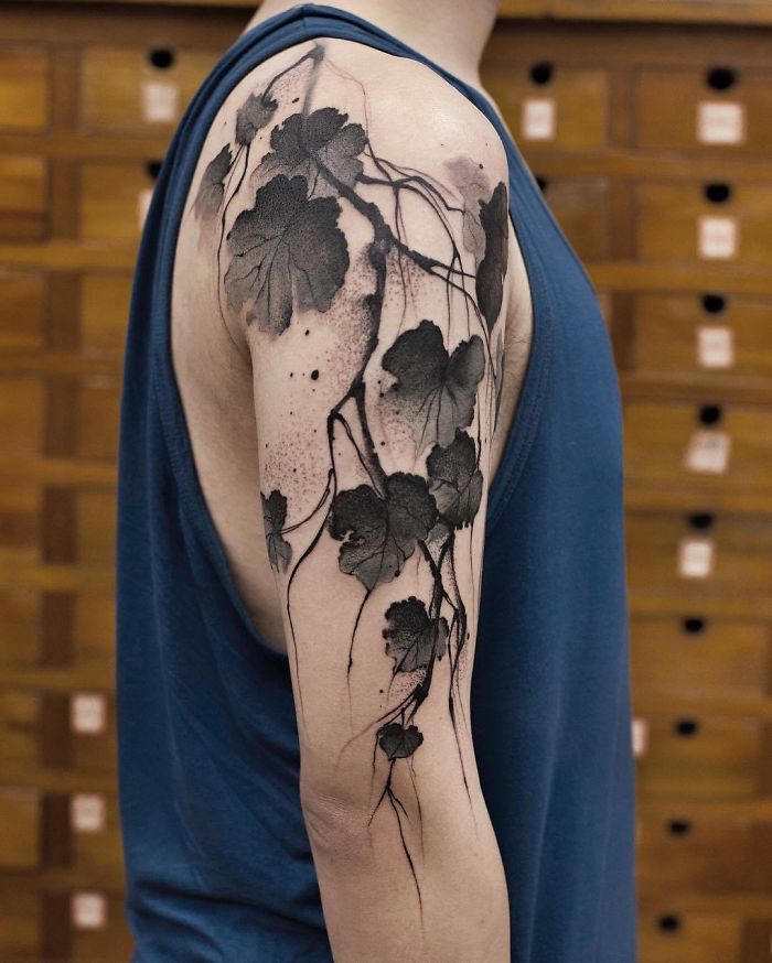 Watercolor tattoo by Chen Jie on arm, featuring black leaves and vines on a person's shoulder in a blue tank top.