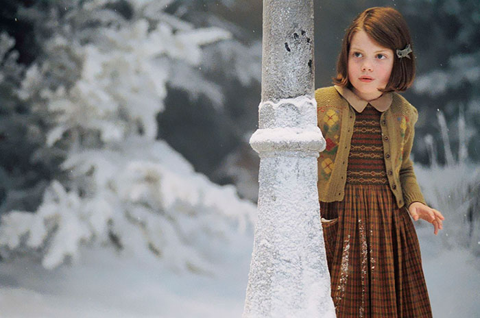 Young girl from The Chronicles Of Narnia movie peeks from behind a snowy lamppost in a winter forest scene.