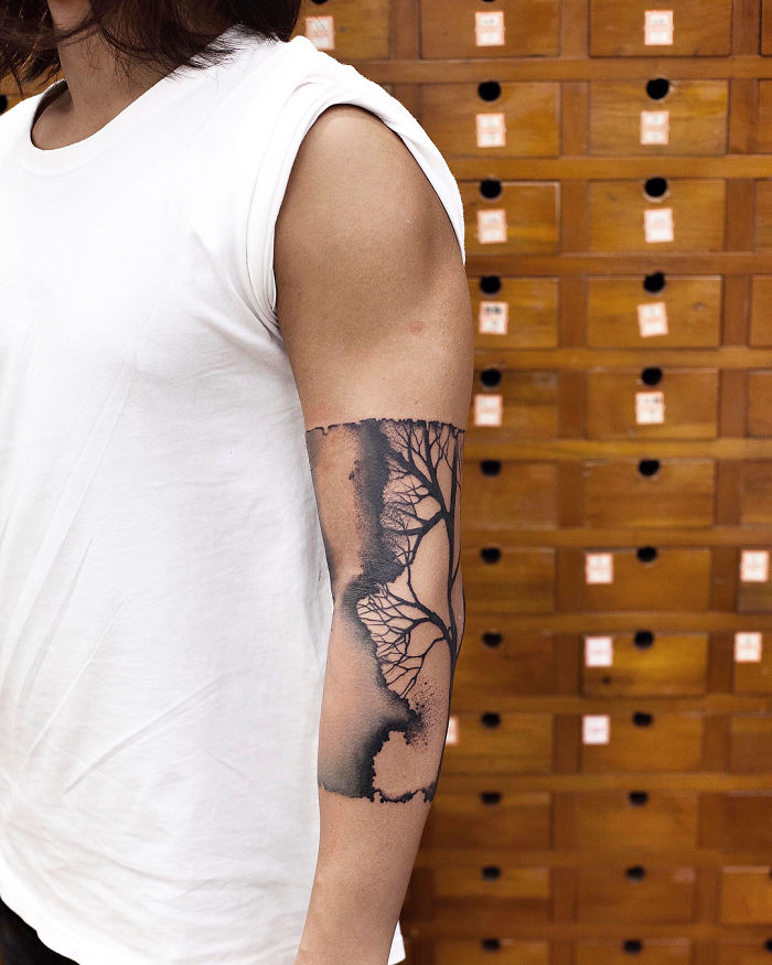 Watercolor tattoo of a tree on a person's arm by artist Chen Jie, against a backdrop of wooden drawers.
