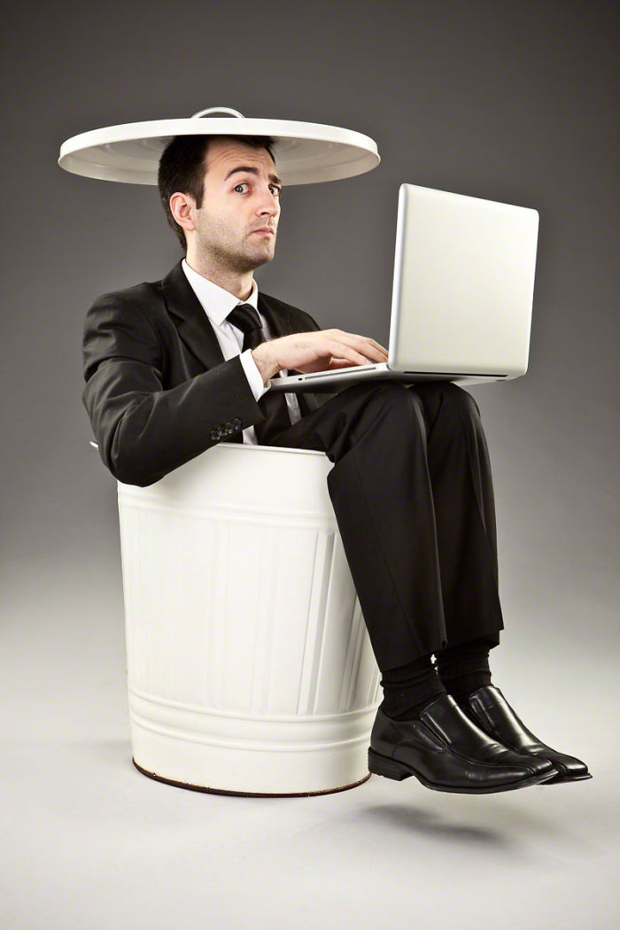 A man in a black suit is sitting in a white metal rubbish bin with a cover on his head like a hat and he is working on a silver laptop