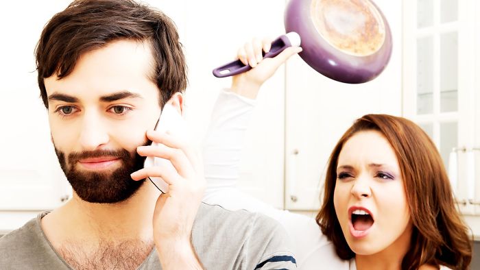 A man is on the phone and an angry woman behind him attempting to hit him with a frying pan