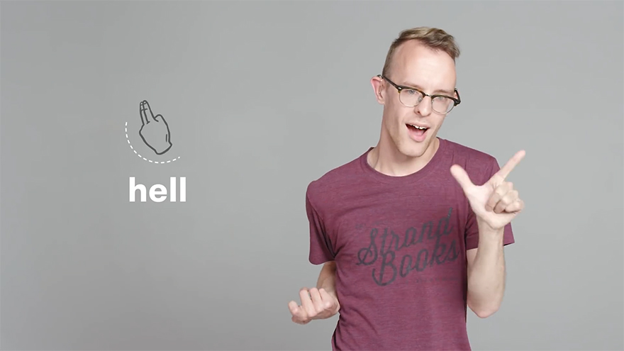Man in glasses demonstrating curse words in sign language, making a gesture for "hell" in a casual setting.