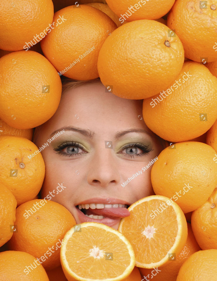 A woman's face among fresh oranges and she is licking one orange that is cut in half