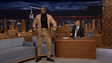 Terry Crews in a tan suit on a talk show set, showcasing his illustration talent. Terry Crews in a tan suit on a talk show set, showcasing his illustration talent.