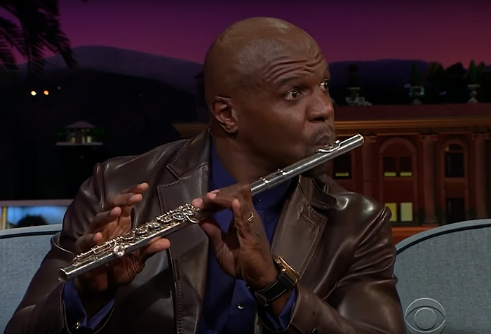 Man playing the flute on a talk show set, wearing a brown leather jacket and dark shirt. Man playing the flute on a talk show set, wearing a brown leather jacket and dark shirt.