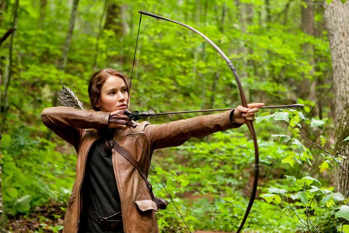 Scene from The Hunger Games movie featuring a character aiming a bow and arrow in a dense forest setting.