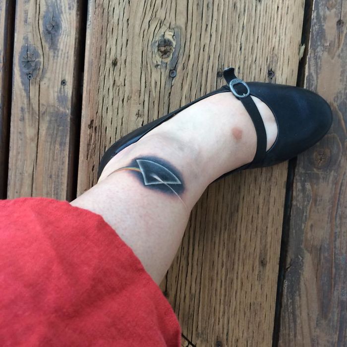 Tattoo on leg with geometric design near ankle, wearing a black shoe on wooden deck.