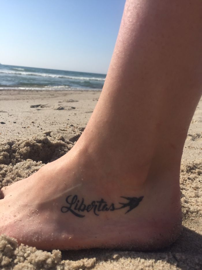 Foot tattoo with "Libertas" and a bird on a sandy beach, highlighting the personal stories behind tattoos.