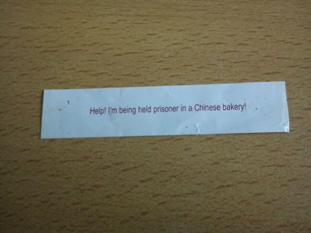 Fortune cookie message on a table reading, "Help! I'm being held prisoner in a Chinese bakery!"